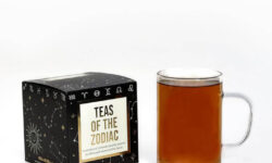 A large square black cardboard box that’s covered in some silver stars with a creamy white label that has Bird & Blend Tea Company Zodiac Collection Cube written in bright gold writing on it next to a short cylindrical clear glass mug full of some dark brown liquid, on a white background.