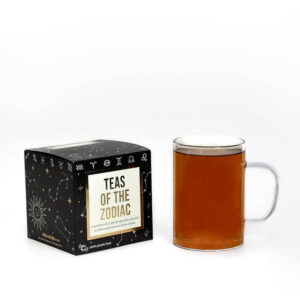 A large square black cardboard box that’s covered in some silver stars with a creamy white label that has Bird & Blend Tea Company Zodiac Collection Cube written in bright gold writing on it next to a short cylindrical clear glass mug full of some dark brown liquid, on a white background.
