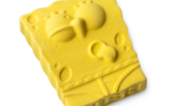 A medium bright yellow rectangular sponge shaped bath bomb that has some large eyes, a sausage nose, and a toothy grin engraved on it, on a white background.