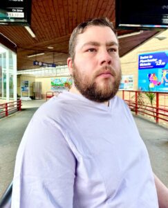 A tall bearded man wearing a light lilac purple v neck t-shirt sat on a silver metal bench.