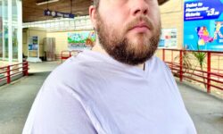 A tall bearded man wearing a light lilac purple v neck t-shirt sat on a silver metal bench.