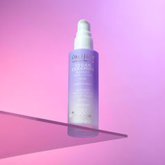 A tall cylindrical clear Glass Bottle full of some pale purple liquid with a silver metal pump lid and a white label that has Pacifica Vegan Ceramide Barrier Face Lotion written in bold black writing on it, on a white background.