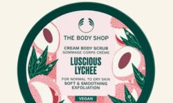 A short cylindrical dark green plastic tub with a dark green plastic lid and a bright pink label that has The Body Shop Luscious Lychee Body Scrub written in dark green writing and some pictures of some bright pink slices of Lychee on it, on a white background.