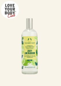 A tall cylindrical clear glass bottle full of some clear liquid with a silver metal lid and a bright yellow label that has The Body Shop Zesty Lime Blossom Hydrating Face & Body Mist written in dark green writing, some pictures of some dark green slices of Lime, and some pictures of some bright white lime blossoms on it, on a white background.