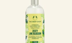 A tall cylindrical clear glass bottle full of some clear liquid with a silver metal lid and a bright yellow label that has The Body Shop Zesty Lime Blossom Hydrating Face & Body Mist written in dark green writing, some pictures of some dark green slices of Lime, and some pictures of some bright white lime blossoms on it, on a white background.