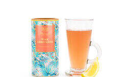 A tall cylindrical bright white cardboard tube with a bright gold plastic lid that has Whittard of Chelsea Pink Lemonade Instant Tea written in bright gold writing and some pictures of some vines on it next to a tall cylindrical clear glass full of some translucent pink liquid with a bright yellow slice of lemon coming out of the top of it, on a white background.