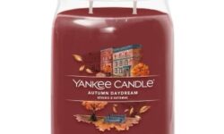 A tall cylindrical glass jar full of some dark red wax with a label that has Yankee Candle written in white writing, Autumn Daydream written in white writing, and a picture of some dark brown trees that are losing their leaves on it, on a white background.