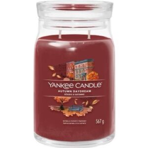A tall cylindrical glass jar full of some dark red wax with a label that has Yankee Candle written in white writing, Autumn Daydream written in white writing, and a picture of some dark brown trees that are losing their leaves on it, on a white background.