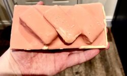 A large rectangular bright orange chocolate bar that has some layers of fluffy golden brown cake, some dark red Raspberry Jam, and some multicoloured sprinkles running through it, on a light wooden worktop.