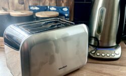 A large rectangular shiny silver metal Toaster that has WeeKett written in bold black writing on it next to a tall cylindrical silver matte plastic kettle with a black matte plastic handle that has WeeKett written in bold black writing on it on a light wooden worktop.