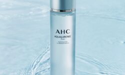 A tall cylindrical white plastic bottle with a white plastic Lid that has AHC Aqualuronic Hydrating Toner written in bold black writing on it, on a white background.
