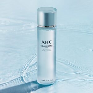 A tall cylindrical white plastic bottle with a white plastic Lid that has AHC Aqualuronic Hydrating Toner written in bold black writing on it, on a white background.