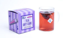 A large square bright pink and purple striped cardboard box with a creamy white label that has Bird & Blend Tea Company x Swizzles Palma Violet Tea Cube written in bright purple writing on it next to a short cylindrical clear glass mug full of some dark red liquid, on a white background.