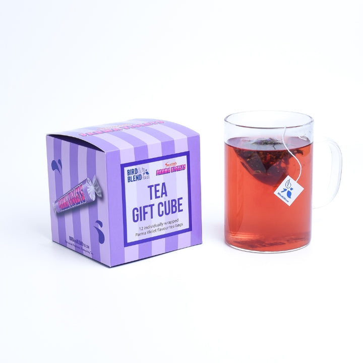 Tea Tastes Terrific | Bird & Blend Tea Company X Swizzles Collection A large square bright pink and purple striped cardboard box with a creamy white label that has Bird & Blend Tea Company x Swizzles Palma Violet Tea Cube written in bright purple writing on it next to a short cylindrical clear glass mug full of some dark red liquid, on a white background.