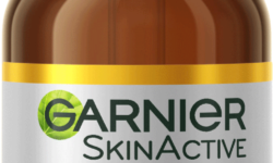 A short rounded cylindrical brown glass bottle with a white rubber pipette lid and a silver label that has Garnier SkinActive Anti-Dark Spot Night Serum written in bold black writing and Vitamin C written in bright orange writing on it, on a white background.