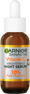 A short rounded cylindrical brown glass bottle with a white rubber pipette lid and a silver label that has Garnier SkinActive Anti-Dark Spot Night Serum written in bold black writing and Vitamin C written in bright orange writing on it, on a white background.