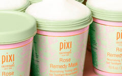 A tall cylindrical white plastic tub with a white plastic lid that has Pixi Rose Remedy Mask written in bright gold writing on it, on a white background.