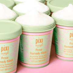 A tall cylindrical white plastic tub with a white plastic lid that has Pixi Rose Remedy Mask written in bright gold writing on it, on a white background.