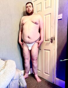 A tall bearded man wearing a black g-string with a white waistband that has Blanc Menswear written in bold black writing on it stood against a white wooden door.