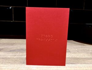 A small dark Red A5 Greetings Card that has Merry Christmas written in Braille Dots on it, on a white background.