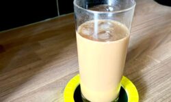 A tall cylindrical glass full of some translucent dark brown liquid and several cubes of ice on top of a large circular bright yellow plastic coaster that has a black plastic ring around it, on a white background.