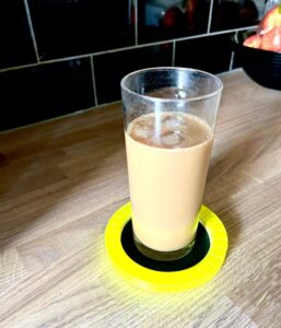 A tall cylindrical glass full of some translucent dark brown liquid and several cubes of ice on top of a large circular bright yellow plastic coaster that has a black plastic ring around it, on a white background.