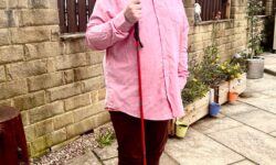 A tall bearded man wearing a pair of rectangular silver metal sunglasses with some dark brown lenses, a bright pink long sleeve shirt, a bright burnt orange v-neck t-shirt, a pair of dark burgundy cord trousers and a pair of shiny red shoes holding a bright red cane stood in front of a dark wooden fence.