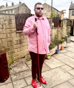 A tall bearded man wearing a pair of rectangular silver metal sunglasses with some dark brown lenses, a bright pink long sleeve shirt, a bright burnt orange v-neck t-shirt, a pair of dark burgundy cord trousers and a pair of shiny red shoes holding a bright red cane stood in front of a dark wooden fence.