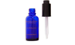 A short cylindrical clear glass bottle full of some dark blue liquid with a black Plastic Pipette Lid and a white label that has Revolution Skincare Overnight Recovery Concentrate written in bright gold writing on it, on a white backhground.