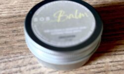 A short cylindrical silver metal tin with a silver metal lid and a white label that has SOS Balm Unscented Balm written in bold black writing on it, on a white background.