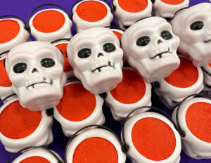 A pile of some small bright white skull shaped bath bombs that all have some black eyes engraved on them, on a white background.