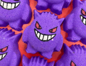 A medium dark purple monster's face shaped Bath Bomb that has some dark Red Eyes engraved on it, on a white background.