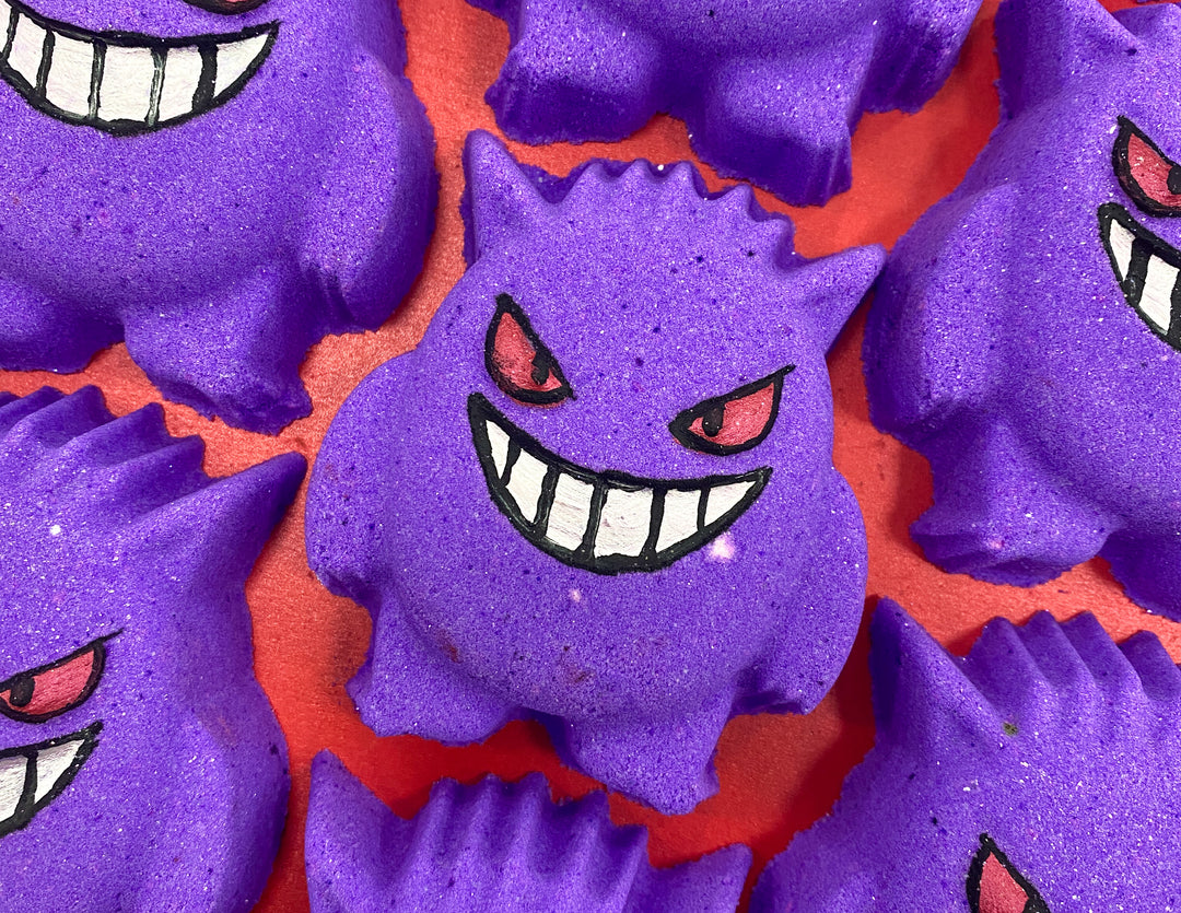 A medium dark purple monster's face shaped Bath Bomb that has some dark Red Eyes engraved on it, on a white background.
