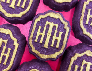 A large bright purple circular shield shaped Bath Bomb that has H.T.H. written in bright gold writing on it, on a white background.