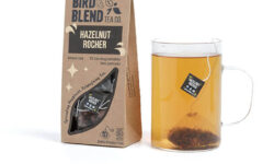 A large rectangular cardboard box that has Bird & Blend Tea Company Hazelnut Rocher Teabag Gift written in bold black writing and a cut out of a large rectangle on it next to a short cylindrical clear glass mug full of some dark brown liquid, on a white background.