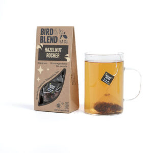 A large rectangular cardboard box that has Bird & Blend Tea Company Hazelnut Rocher Teabag Gift written in bold black writing and a cut out of a large rectangle on it next to a short cylindrical clear glass mug full of some dark brown liquid, on a white background.