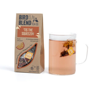 A large rectangular cardboard box that has Bird & Blend Tea Company Tis The Squeezon Teabag Gift written in bold black writing and a cut out of a large slice of Orange on it next to a short cylindrical clear glass mug full of some dark brown liquid , on a white background.