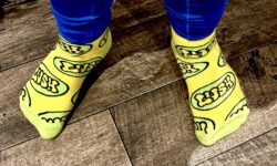 A tall man wearing a pair of shiny dark blue leggings and a pair of bright yellow socks with some bright lime green toes and heels that have a lime green oval pattern and Lush written in bright yellow writing all over them stood on a light wooden laminate floor.