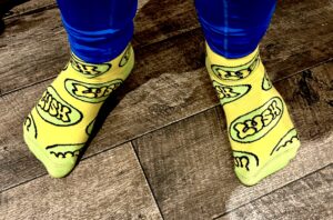 A tall man wearing a pair of shiny dark blue leggings and a pair of bright yellow socks with some bright lime green toes and heels that have a lime green oval pattern and Lush written in bright yellow writing all over them stood on a light wooden laminate floor.
