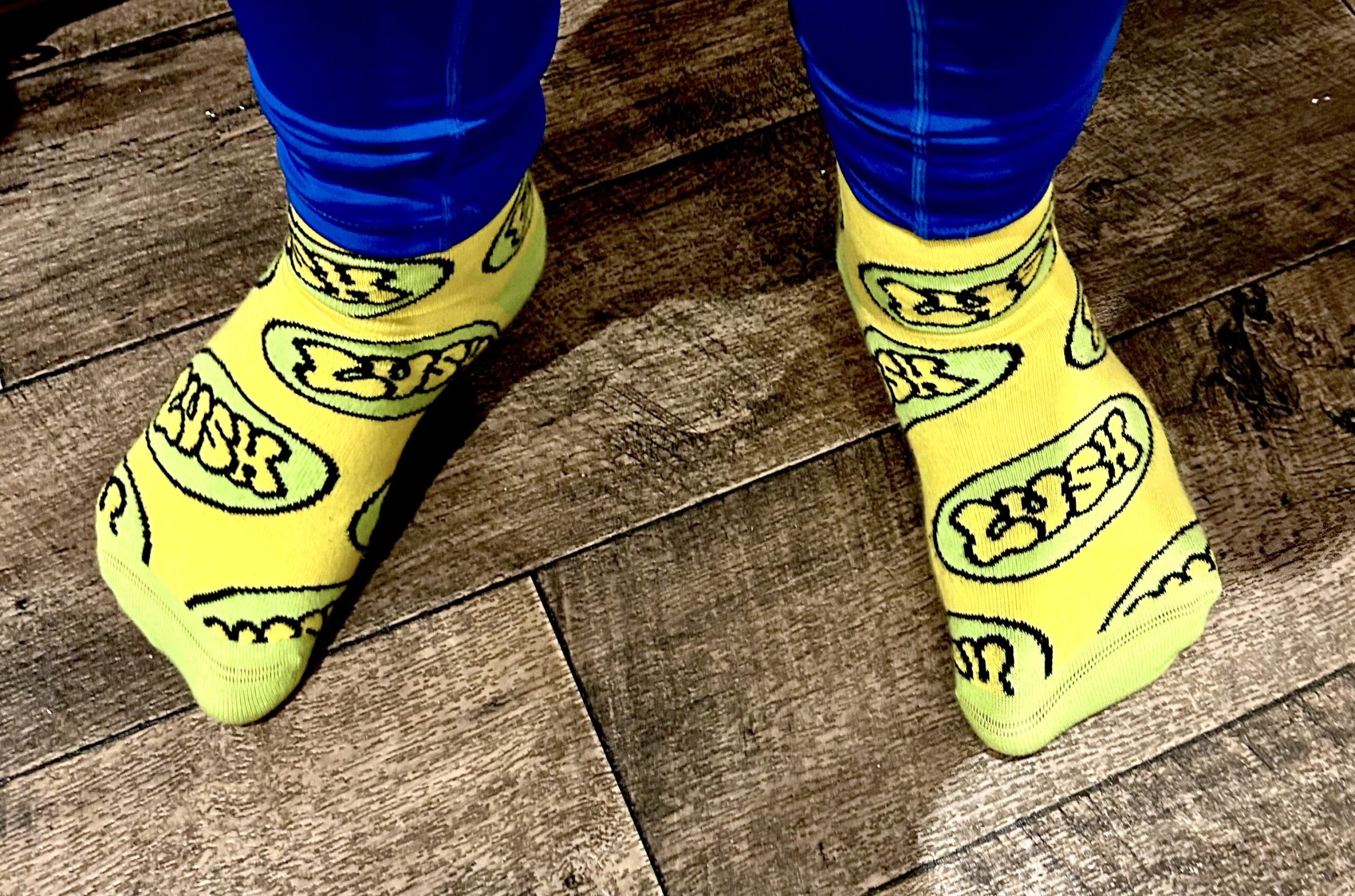 A tall man wearing a pair of shiny dark blue leggings and a pair of bright yellow socks with some bright lime green toes and heels that have a lime green oval pattern and Lush written in bright yellow writing all over them stood on a light wooden laminate floor.