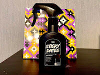A tall black plastic Kitchen spray bottle that has Lush Sticky Dates Body Spray written in bright white writing on it on a dark wooden cabinet.