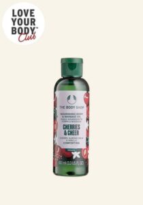 A short and thin cylindrical dark green plastic bottle with a dark green plastic lid and a bright pink label that has The Body Shop Cherries & Cheer Body & Massage Oil written in dark green writing and a picture of some bright red Cherries on it, on a white background.