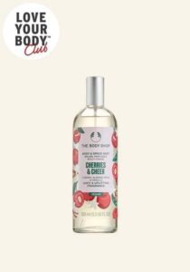 A tall cylindrical clear glass bottle full of some clear liquid with a silver metal lid and a bright pink label that has The Body Shop Cherries & Cheer Body & Space Mist written in dark green writing and a picture of some bright Red Cherries on it, on a white background.