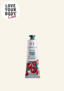 A long and thin conical metallic white tube with a metallic dark green lid and a bright pink label that has The Body Shop Cherries & Cheer Hand Balm written in dark green writing and some pictures of some bright red cherries on it, on a white background.
