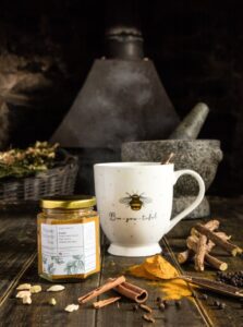 A short hexagonal glass jar full of some bright orange powder next to a tall cylindrical white cup that has a Bee engraved on it, a small cylindrical dark grey stone Pestle and Mortar, a pile of some dark orange Turmeric Powder and some dark brown Cinnamon Sticks on a dark wooden table, on a white background.