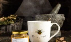 A short hexagonal glass jar full of some bright orange powder next to a tall cylindrical white cup that has a Bee engraved on it, a small cylindrical dark grey stone Pestle and Mortar, a pile of some dark orange Turmeric Powder and some dark brown Cinnamon Sticks on a dark wooden table, on a white background.