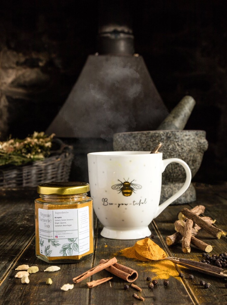 Tea Tastes Terrific | Divine Teas Organic Turmeric Tea 15g Jar | Gifted A short hexagonal glass jar full of some bright orange powder next to a tall cylindrical white cup that has a Bee engraved on it, a small cylindrical dark grey stone Pestle and Mortar, a pile of some dark orange Turmeric Powder and some dark brown Cinnamon Sticks on a dark wooden table, on a white background.