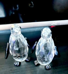 Two small black and white crystal penguins in front of a large rectangular black flatscreen Tv, on a black wooden cabinet.
