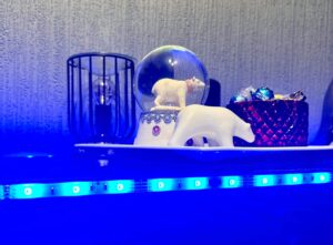 A large bright white spherical snow globe that has a polar bear stood on all four paws inside it next to a small white polar bear that’s stood on all four paws on top of a large rectangular white plate and is surrounded by some bright blue strip lights, on a black wooden cabinet.