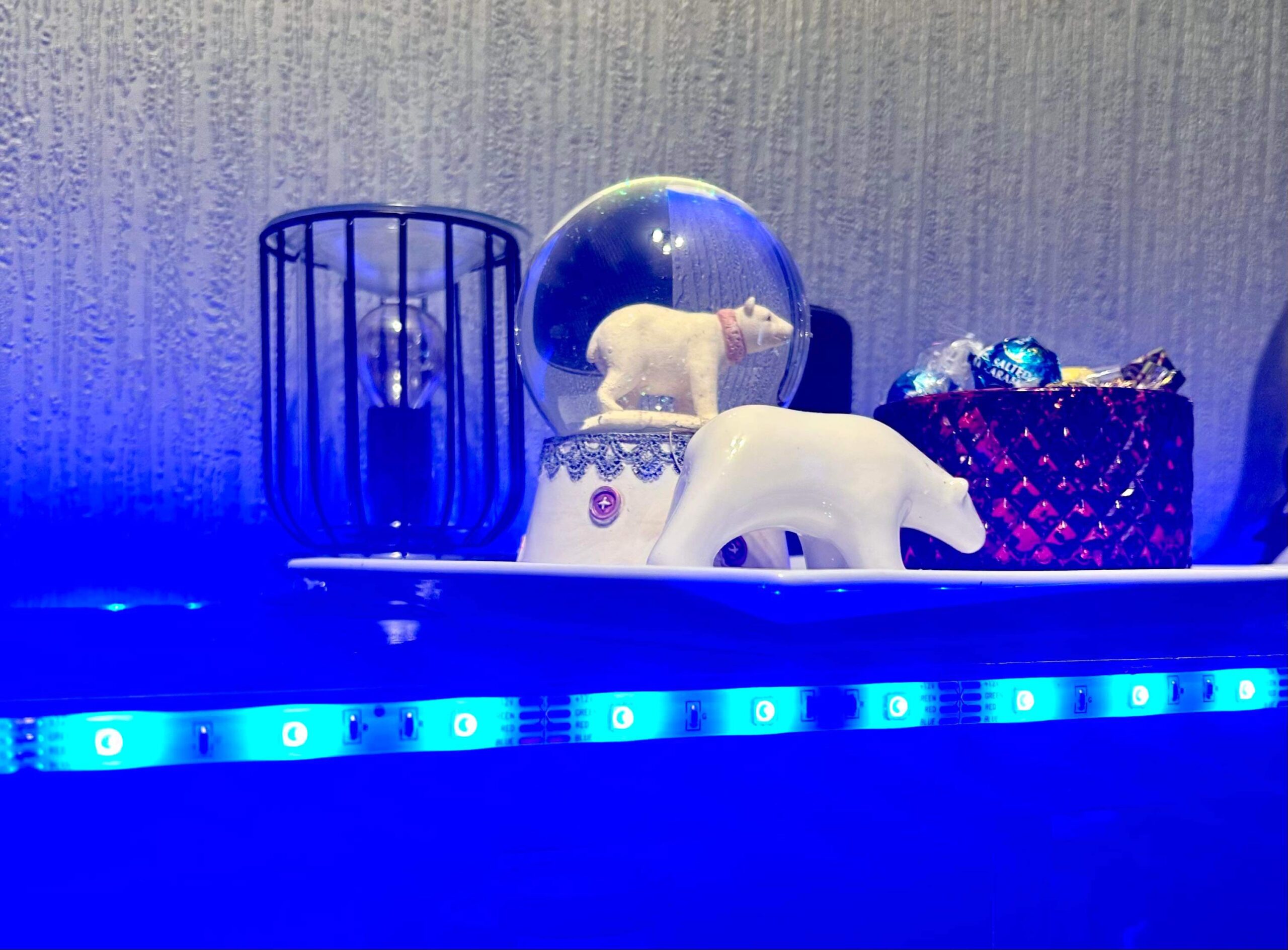 A large bright white spherical snow globe that has a polar bear stood on all four paws inside it next to a small white polar bear that’s stood on all four paws on top of a large rectangular white plate and is surrounded by some bright blue strip lights, on a black wooden cabinet.